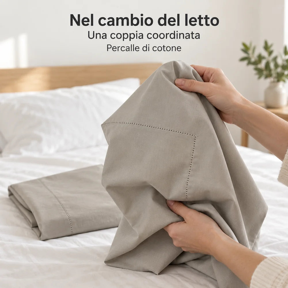 Una federa Greige raccolta fra le mani, la seconda ripiegata sul letto. Foto illustrativa: Greige. Guanciali, arredi e accessori esclusi.
